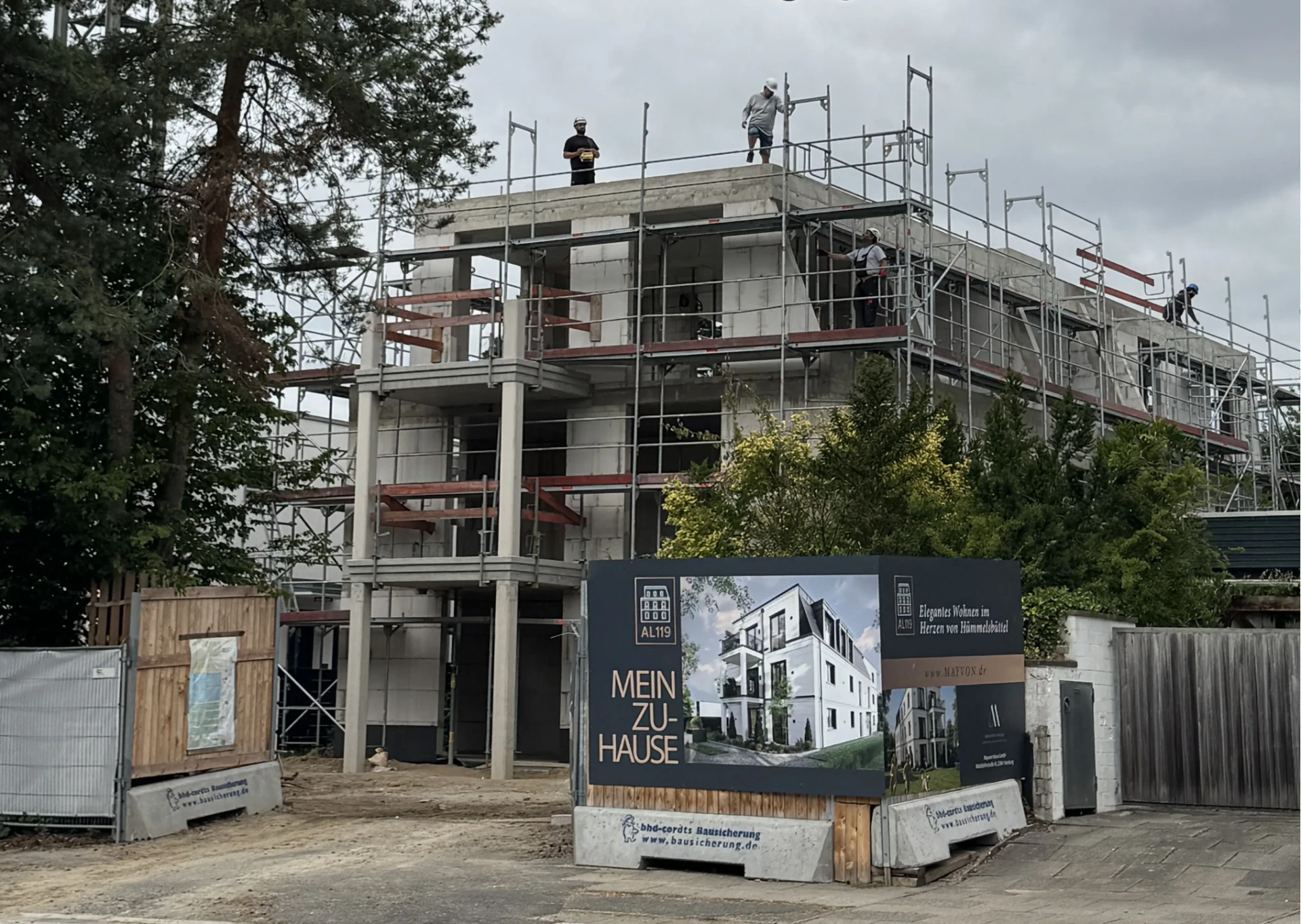 Baustelle Alte Landstraße 119 im August 2026 — Rohbau mit Bauzaunbanner und Arbeitskräften auf dem Dach